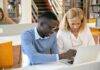 female teacher helping ethnic male student working on laptop in library