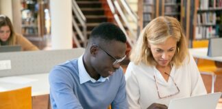 female teacher helping ethnic male student working on laptop in library