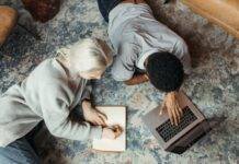 man using a laptop beside a woman reclining on floor carpet and writing on a paper