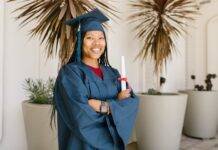 woman in academic gown holding a rolled certificate while smiling at the camera