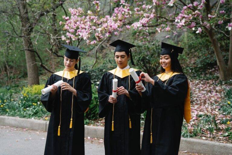 students in graduation gowns and hats unfolding certificates and magnolia in background