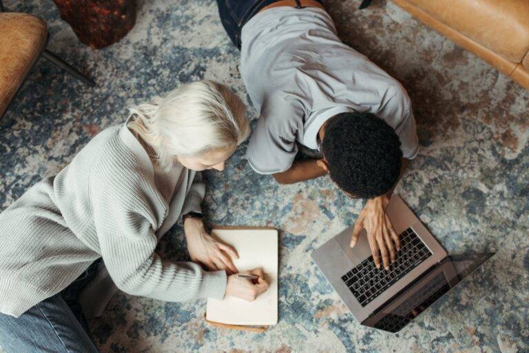 man using a laptop beside a woman reclining on floor carpet and writing on a paper