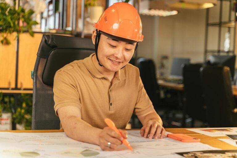 man in brown polo shirt and orange hard hat smiling while writing on paper