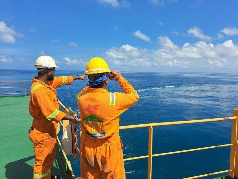 two men in orange safety gear stand on the deck of a ship